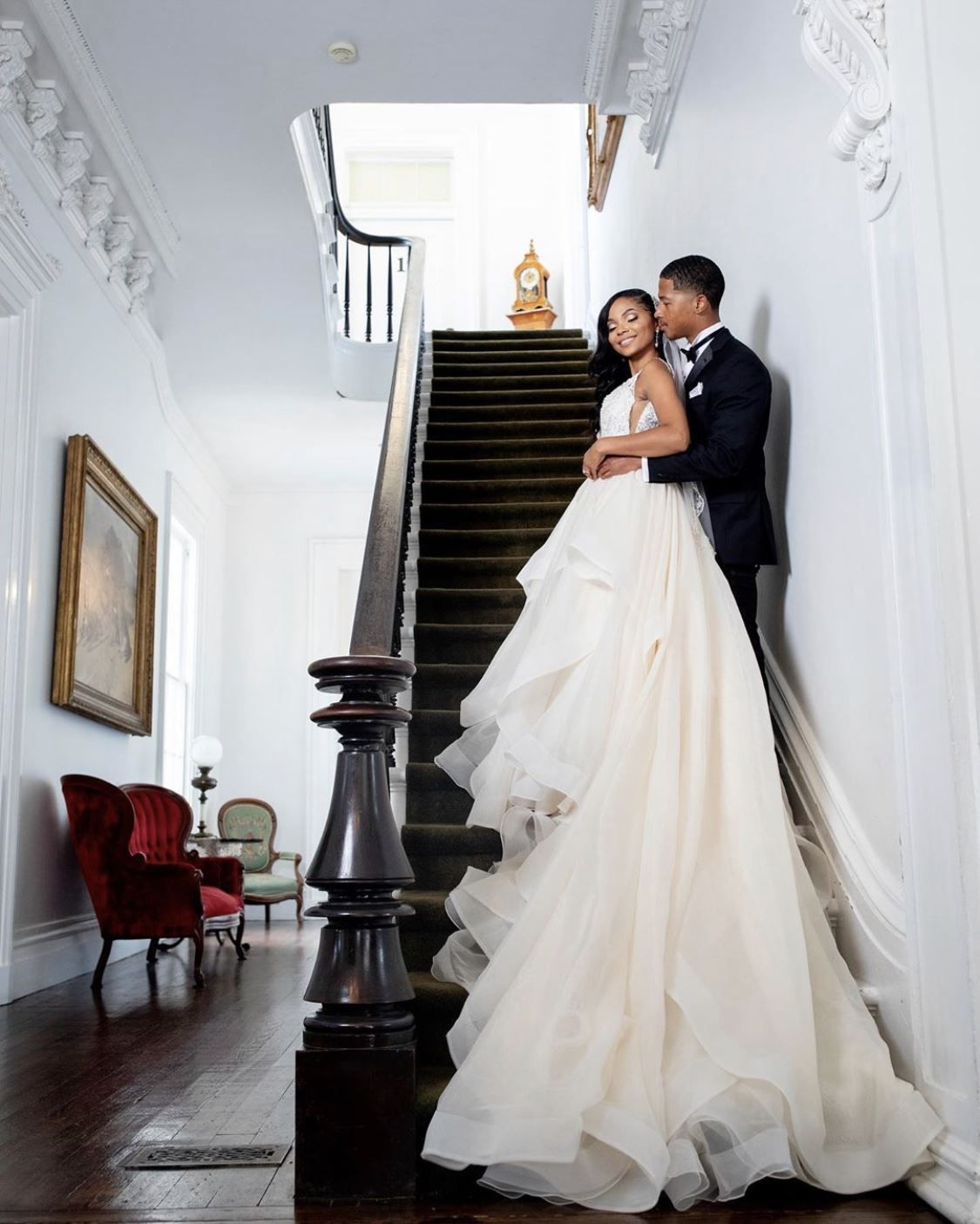Couple embraces on a staircase. Bride wears a flowing dress; groom in a tuxedo. Elegant hallway.