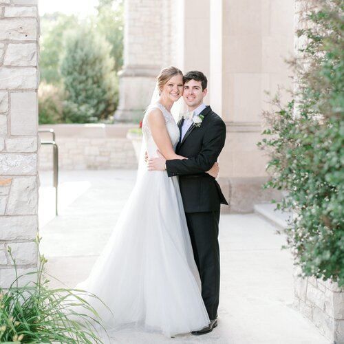 Bride and groom hugging, smiling, outside a building with stone columns. She wears a white gown, he wears a black suit.