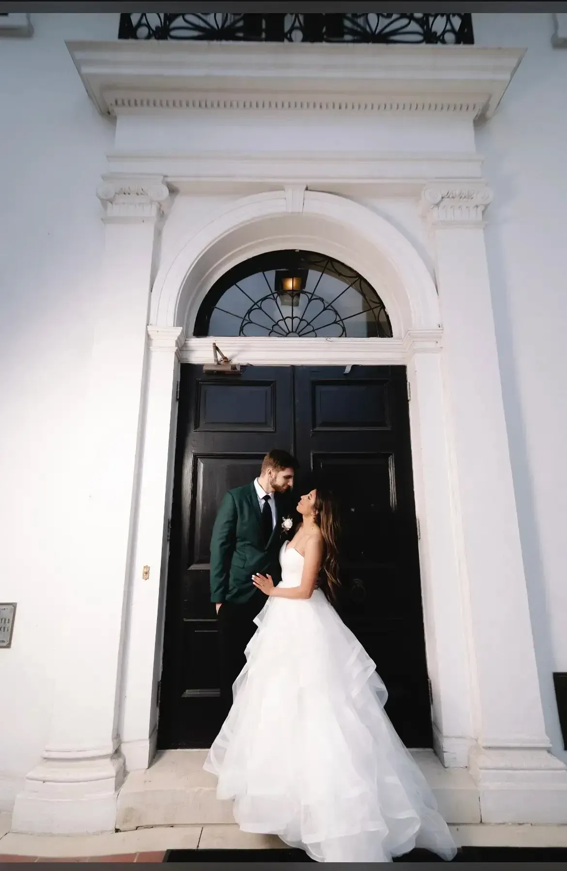 Couple in wedding attire embrace at a black door framed by white architecture.