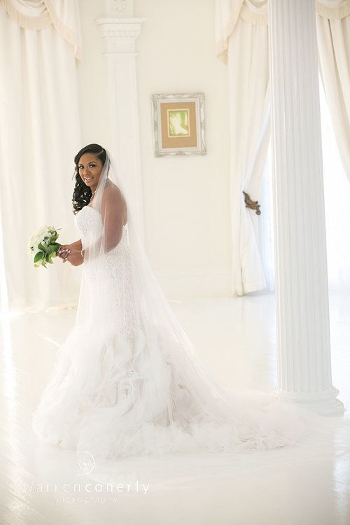 Bride in white wedding dress holding bouquet, inside white room with columns and curtained windows.