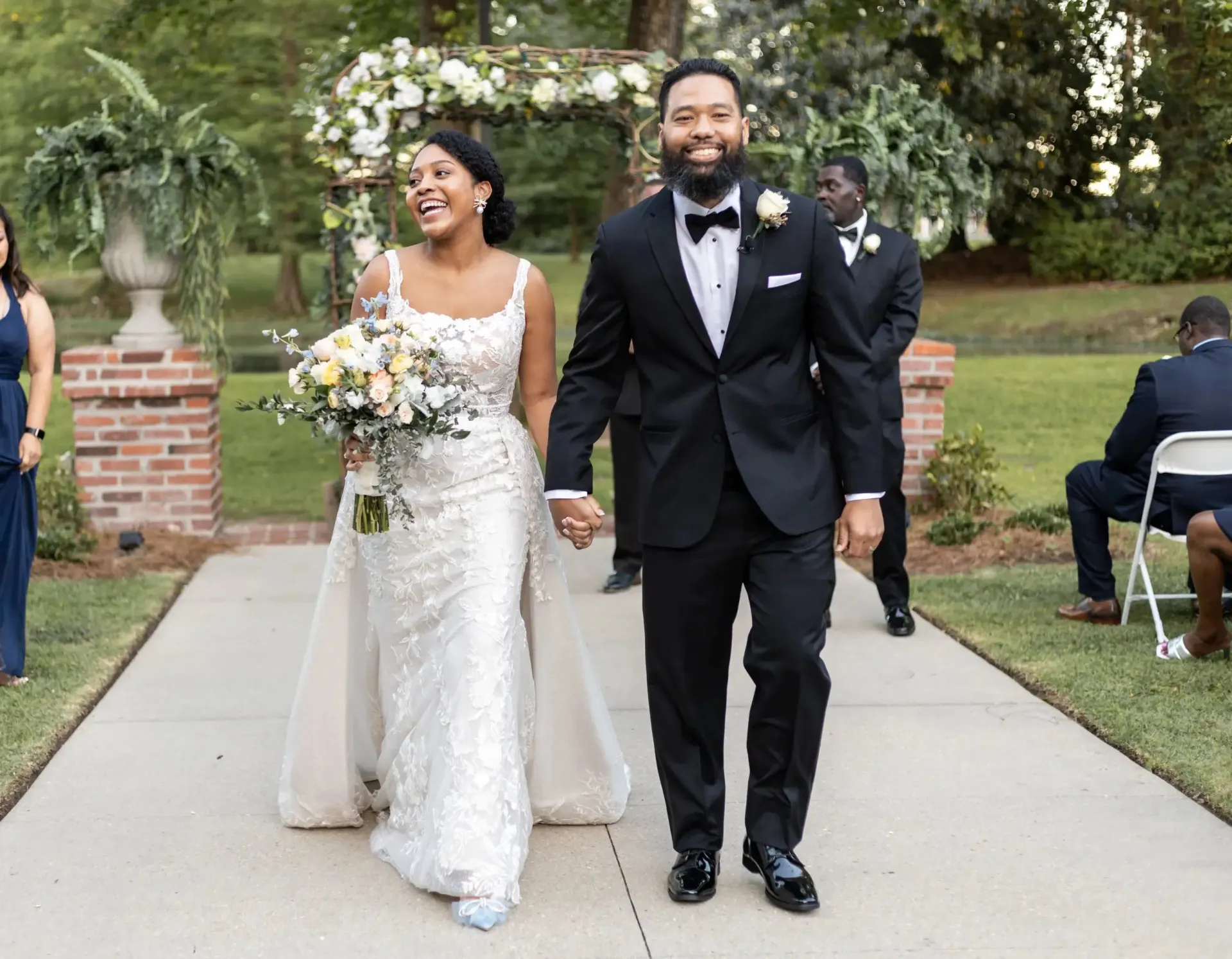 Newlyweds smiling, walking down aisle after wedding. Green grass, floral arch, and a brick pathway.