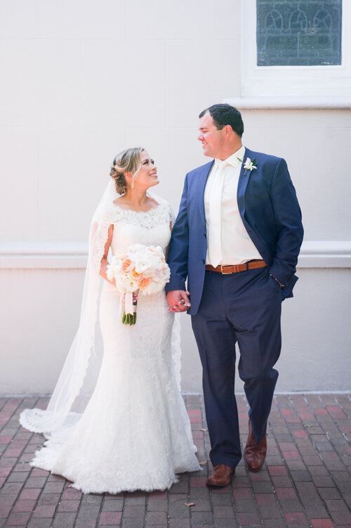 Newlyweds hold hands and walk; bride in lace dress, groom in blue suit.