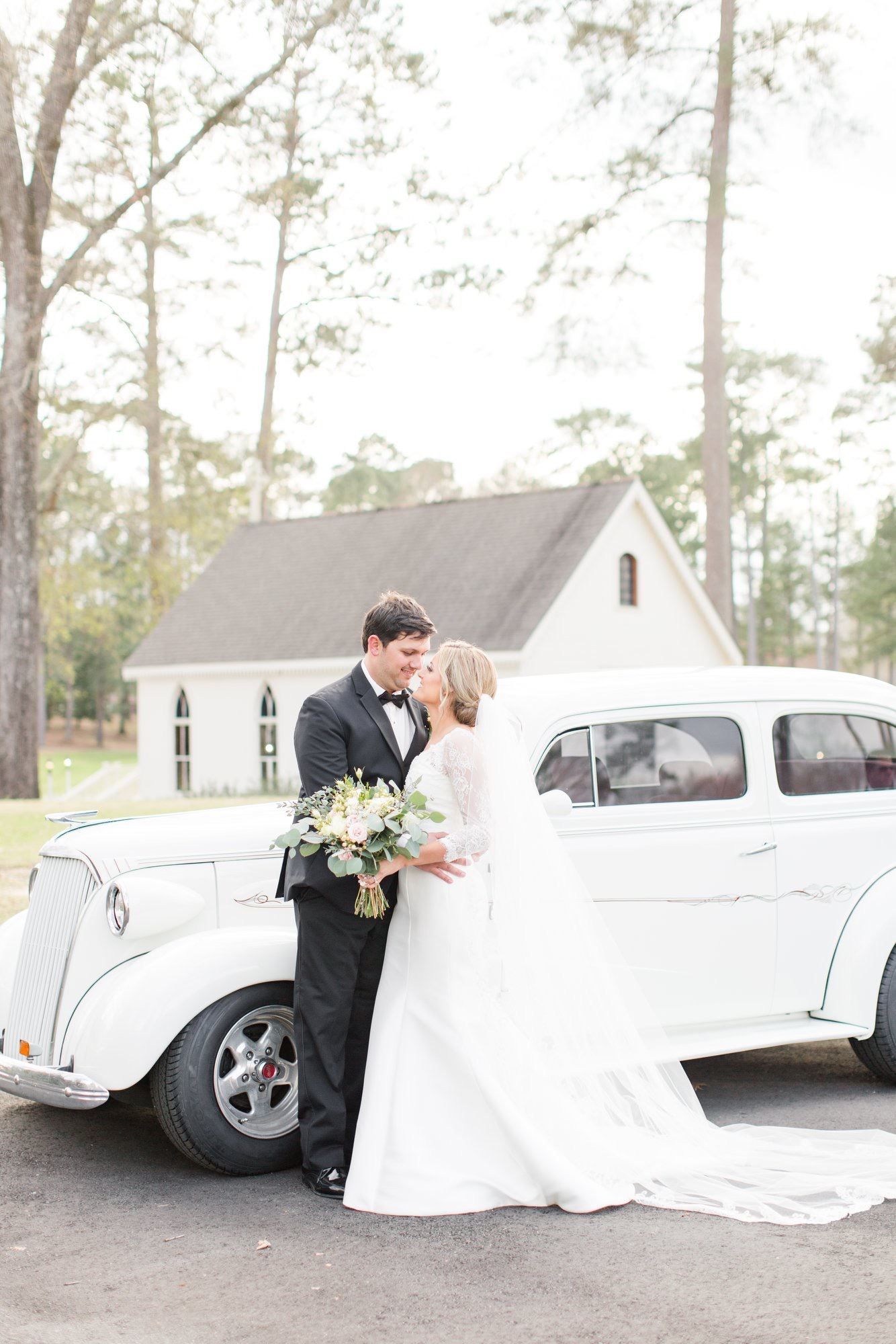 Bride and groom kissing by a white vintage car in front of a white chapel.