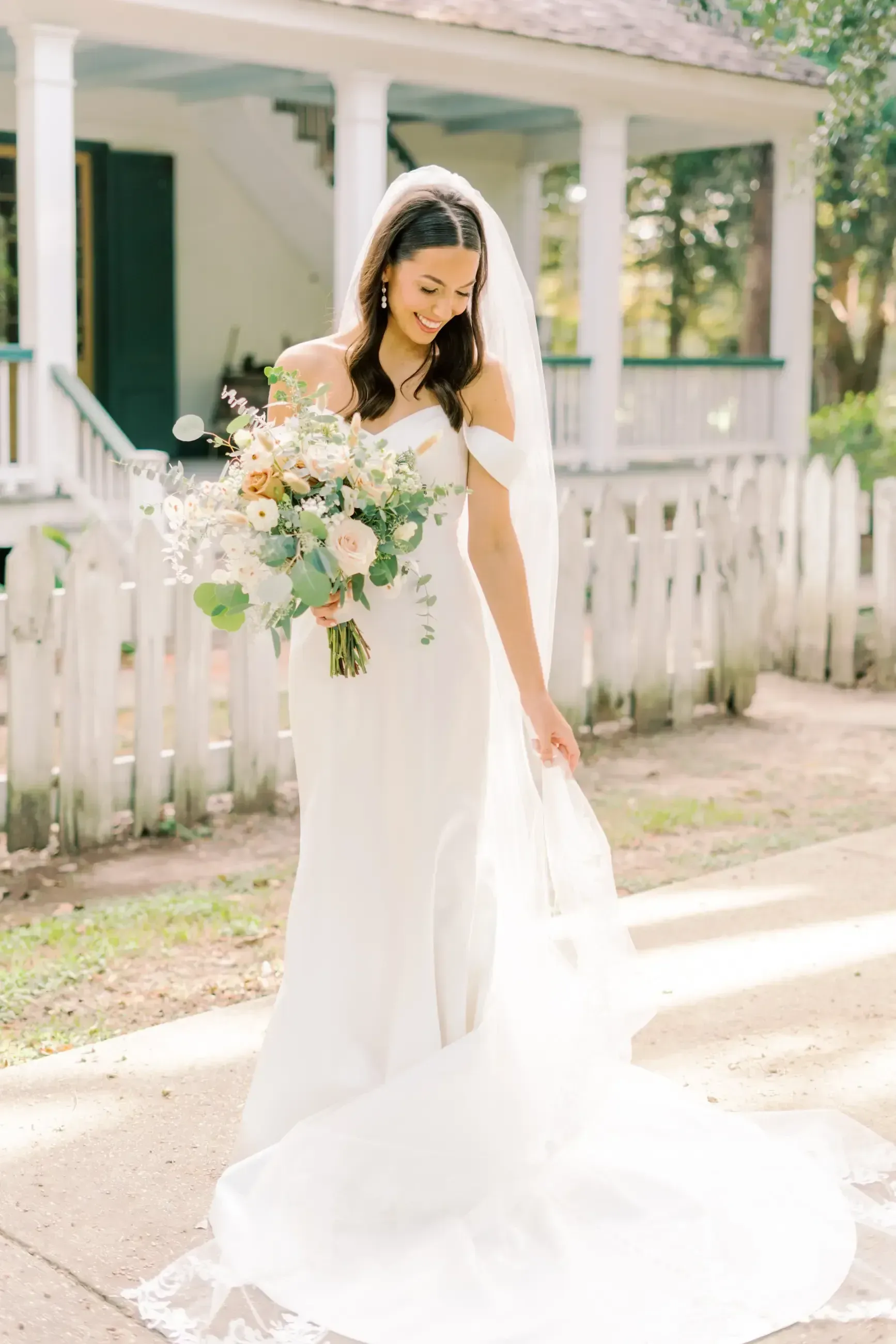 Bride in white gown, smiling, holding bouquet, standing near a white picket fence and house.