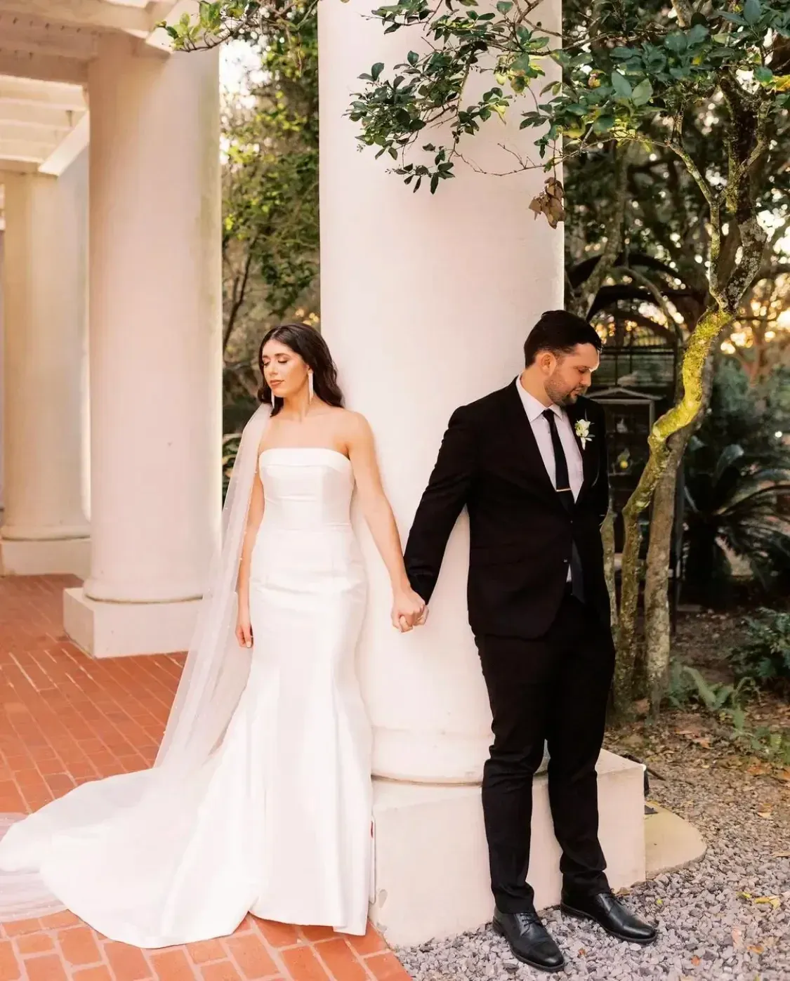 Bride and groom in formal wear holding hands, leaning against columns outdoors.