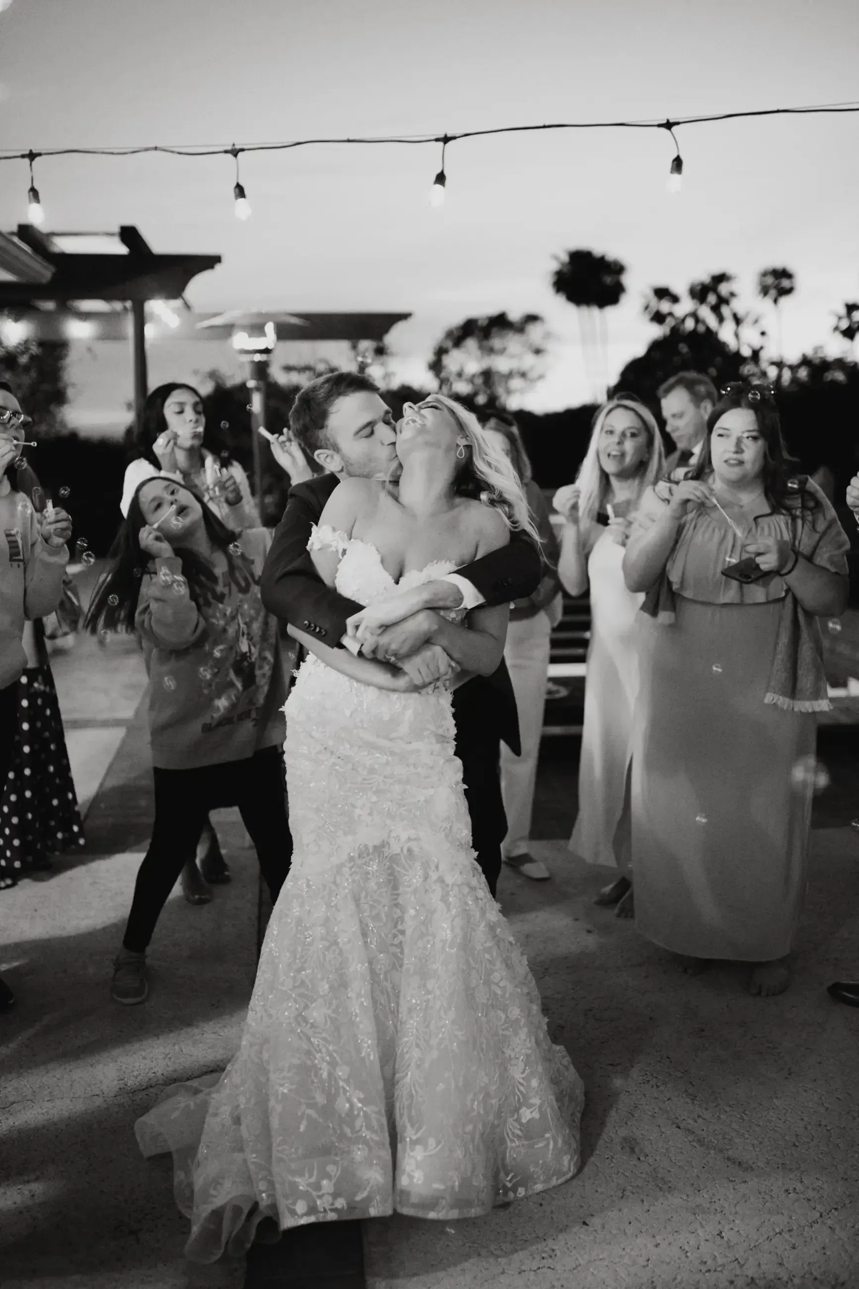 Couple kissing, embraced, at outdoor evening wedding reception, guests cheering, string lights above.