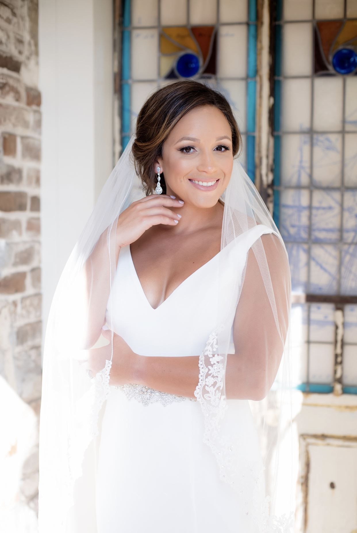 Bride in white wedding dress, veil, and jewelry smiles near stained glass window.