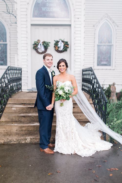 Bride and groom pose on steps in front of a white church. She wears a lace gown; he wears a navy suit.