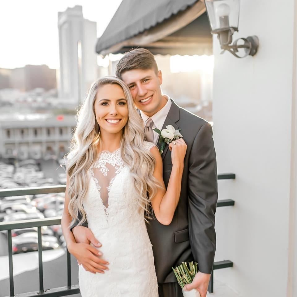 Bride and groom smiling on a balcony; woman in white dress, man in suit. City background.