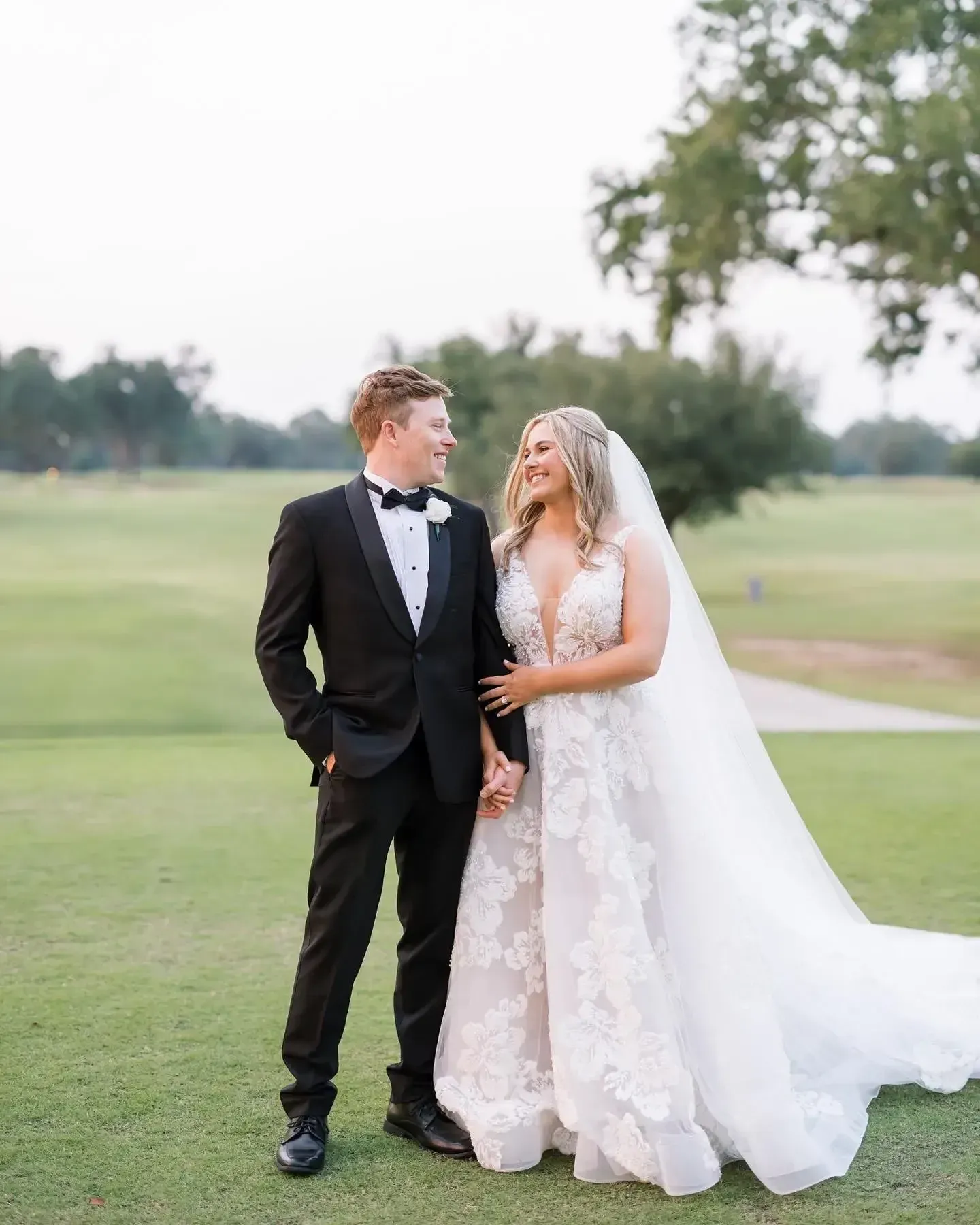 Bride and groom holding hands, smiling at each other on a golf course. She wears a white gown and veil; he wears a tuxedo.