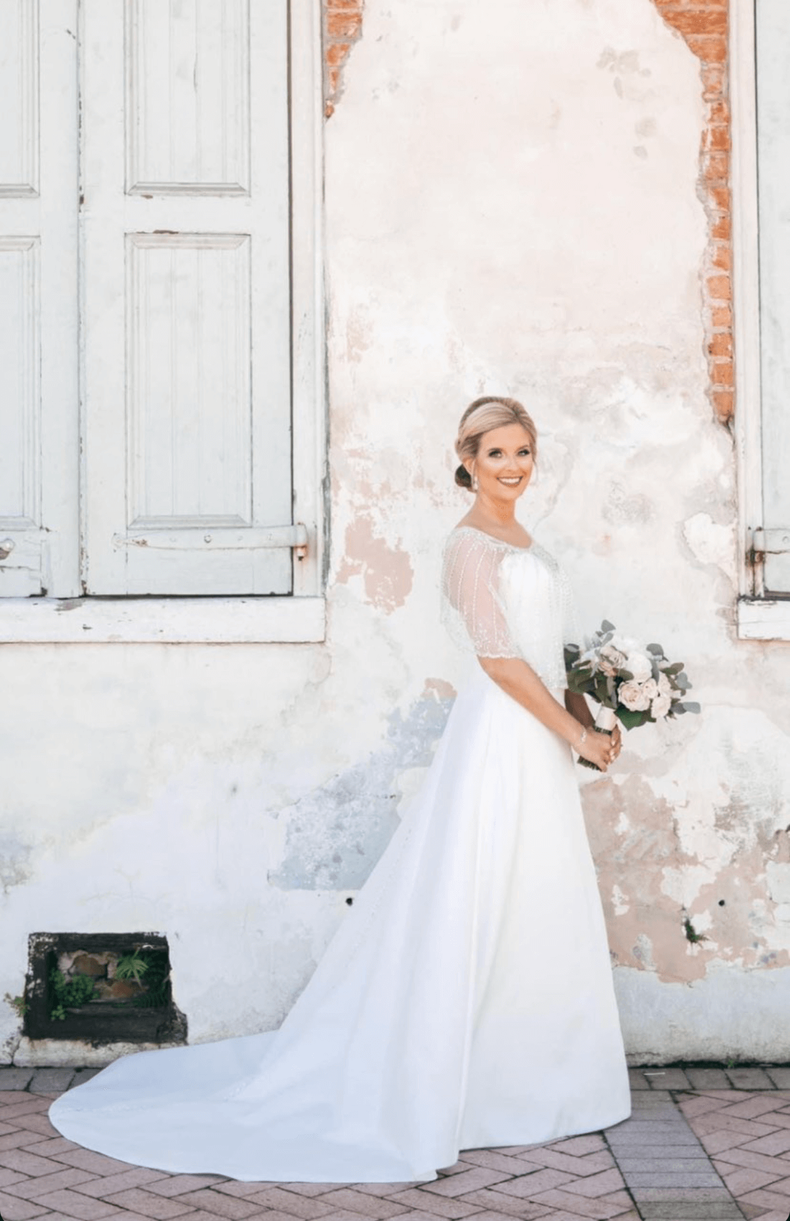 Bride in white dress, holding bouquet, smiles near peeling white wall with shutter.