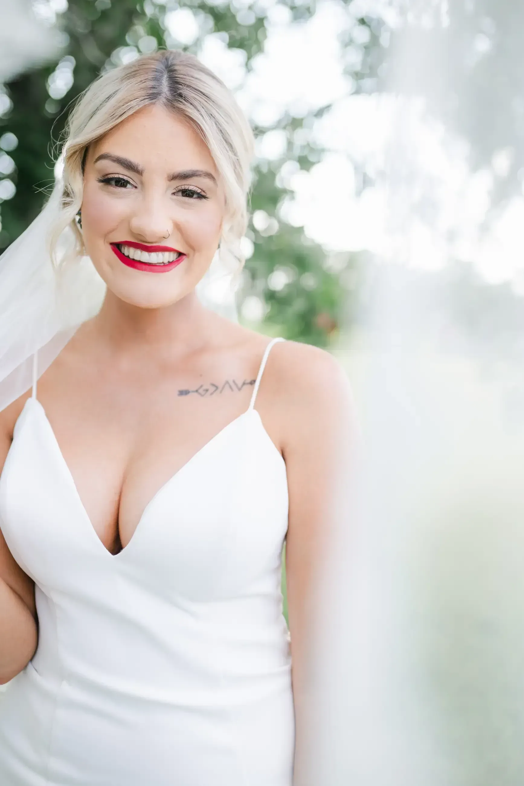 Bride in a white gown smiles with red lipstick, veil, and blonde updo; outdoors.