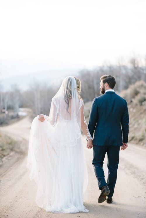 Bride and groom walk hand-in-hand down a dirt road. Bride's dress is white, groom wears a blue suit.