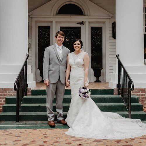 A newly married couple stands on steps. The woman wears a white lace dress and holds flowers, and the man wears a gray suit.