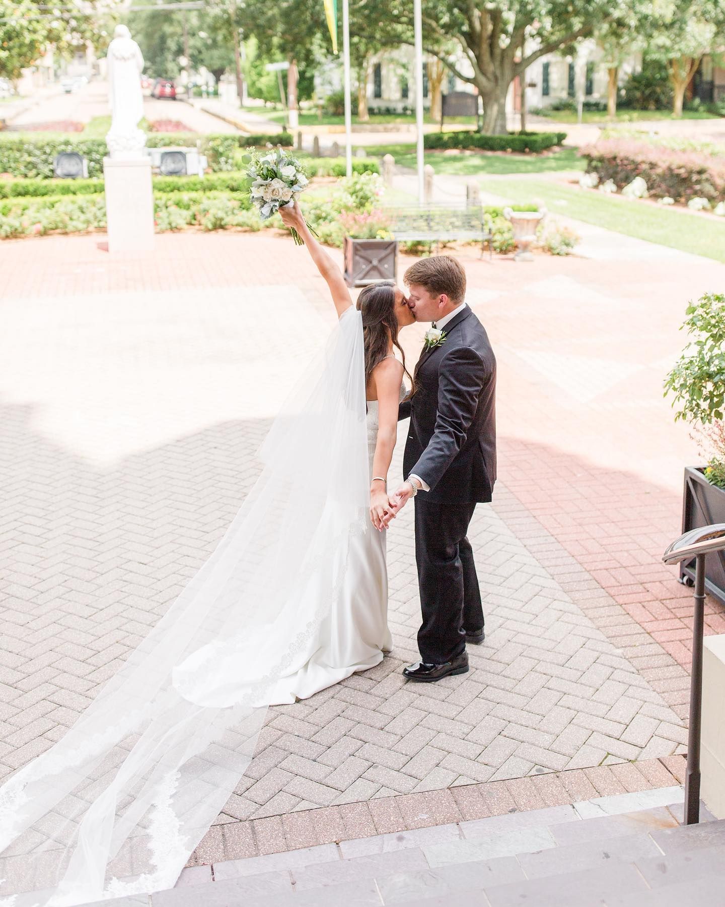 Newlyweds kiss, bride raises bouquet. Long veil, dark suit, brick courtyard, outdoor setting.