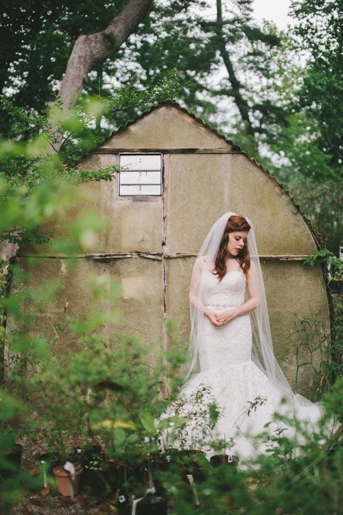 Bride in a white wedding dress and veil stands in front of a weathered shed, surrounded by greenery.
