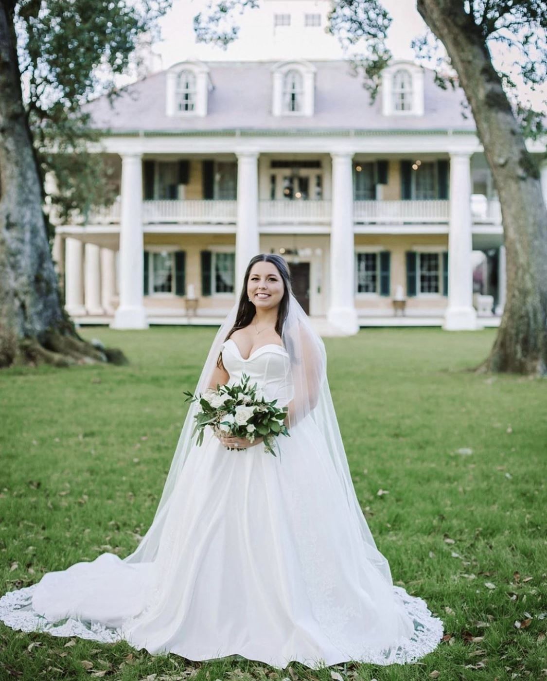 Bride in a white gown and veil holds a bouquet, posing in front of a large white mansion.