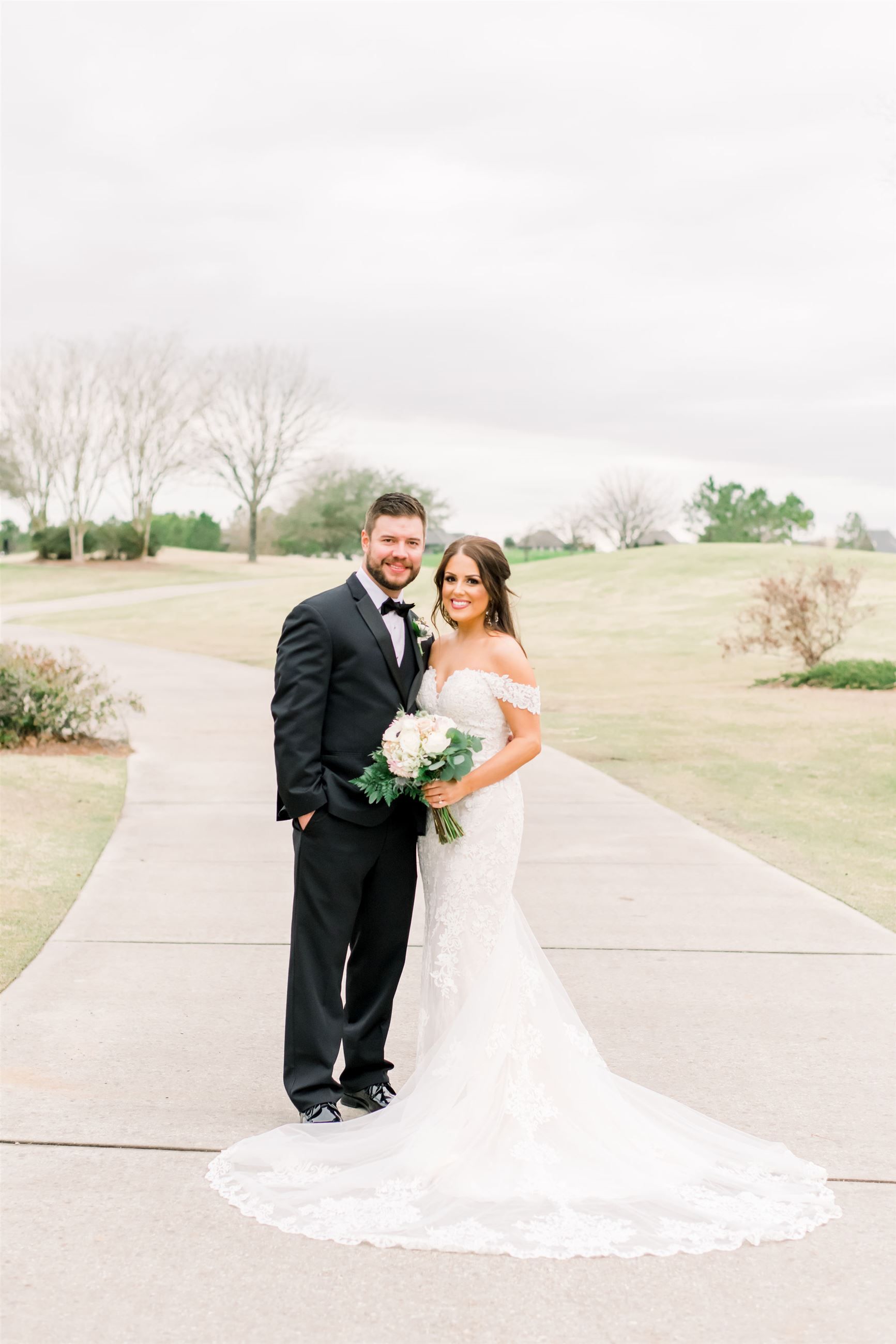 Bride and groom posing on a path. She wears a white lace gown and holds a bouquet; he wears a black tuxedo.