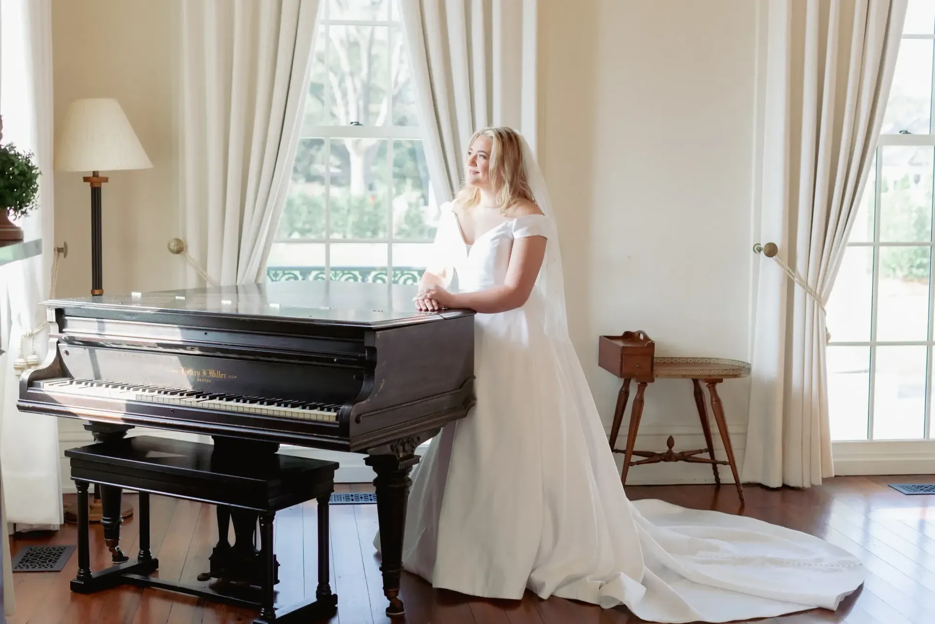 Bride in white gown leans on a grand piano in front of a window.