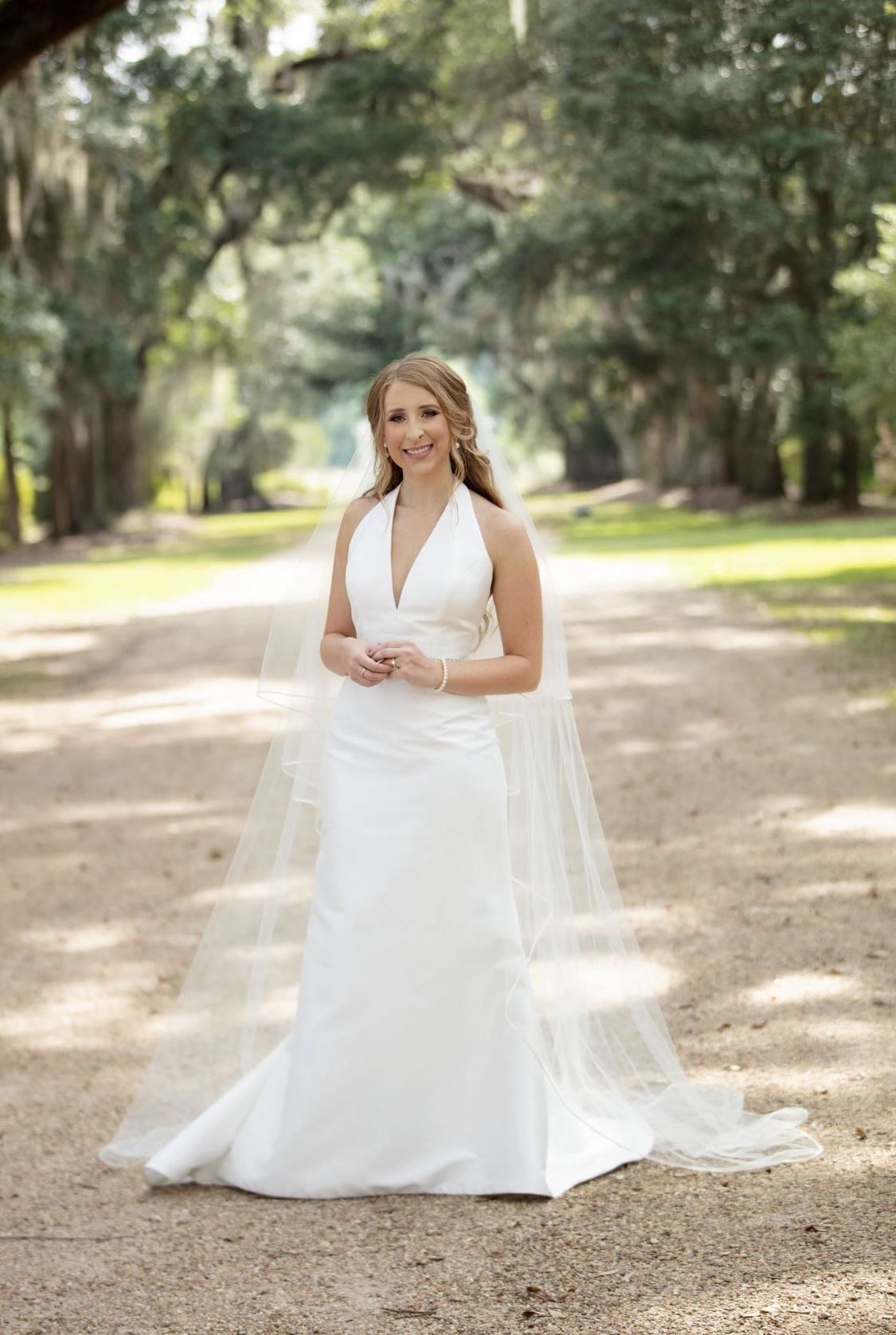 Bride in white halter wedding dress stands on a dirt path under tree canopy.