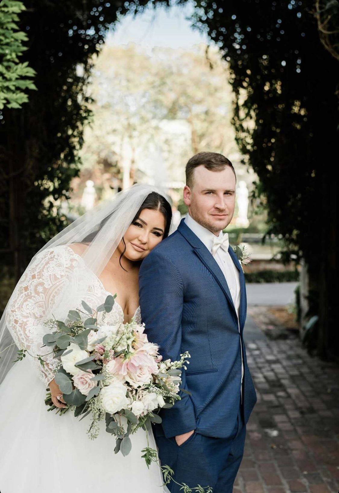 Bride in a white dress leans on groom in blue suit; holding bouquet, smiling. Under a plant archway.