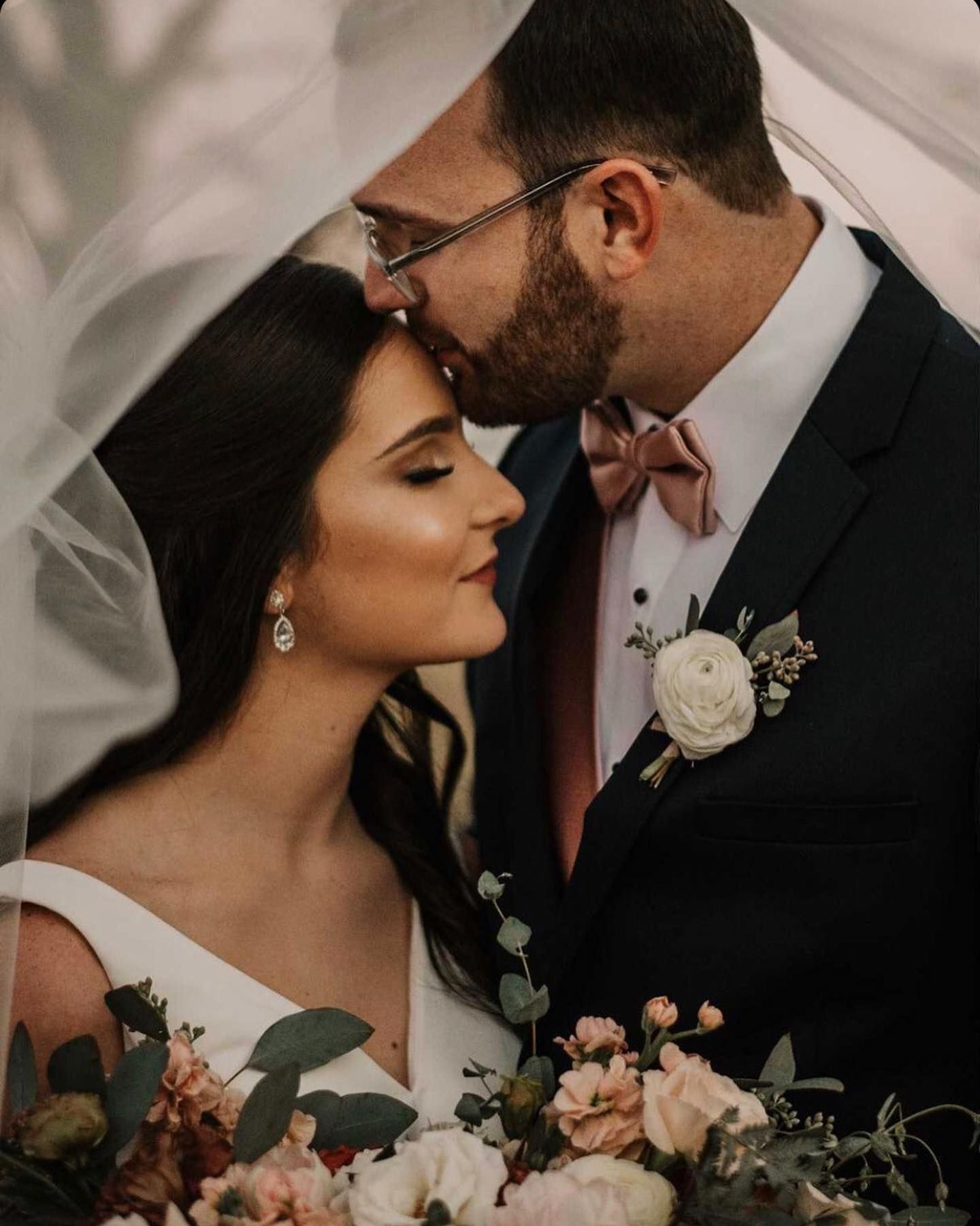 Bride and groom kissing under a veil, holding flowers. She has long dark hair, he wears glasses and a suit.