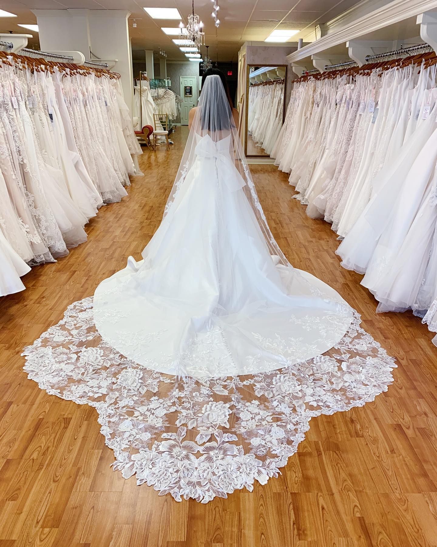 Woman in a wedding dress and veil stands in a bridal shop, surrounded by gowns on display.