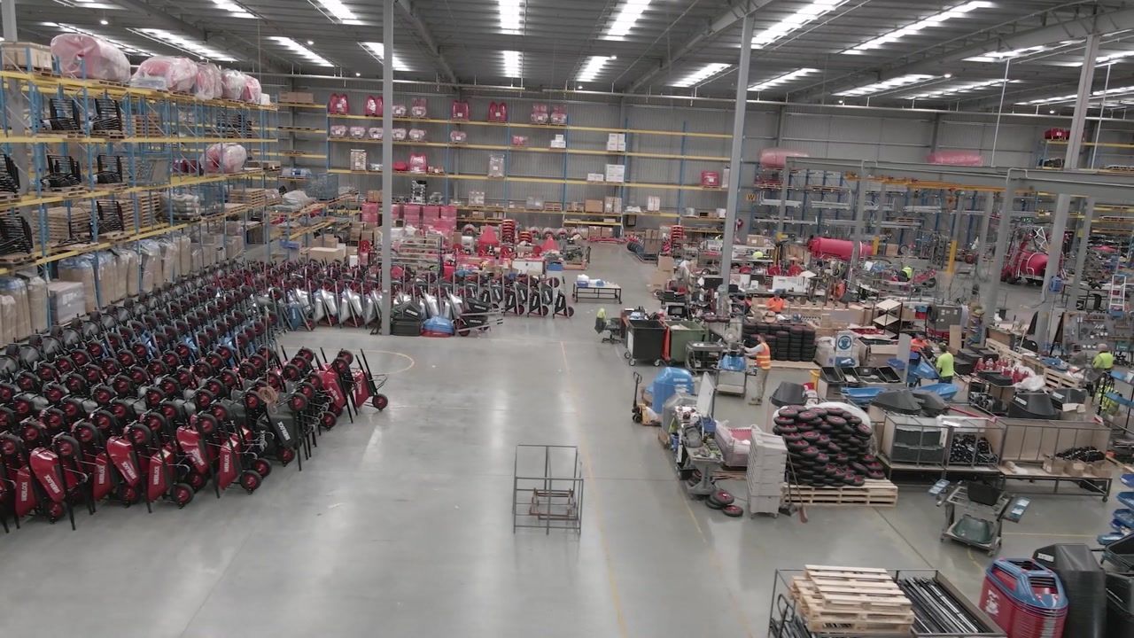 Warehouse interior, golf bags in rows, shelves of goods, workers, pallets, overhead lighting.