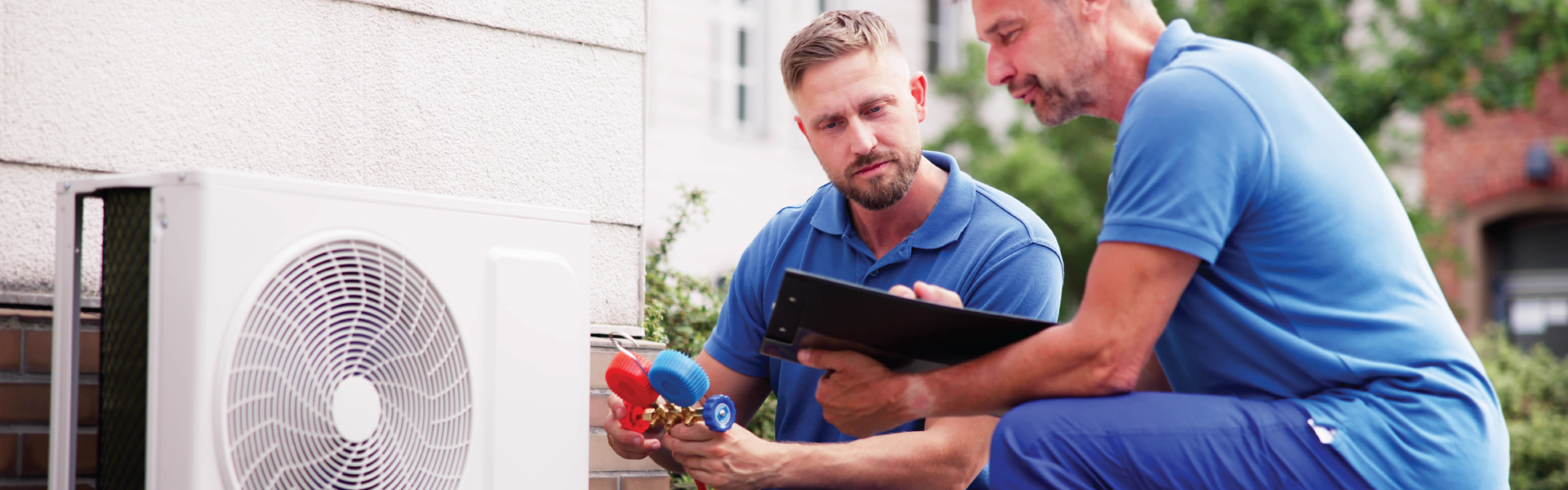 Two technicians examining an air conditioning unit. One holds a gauge while another looks at a clipboard outside.