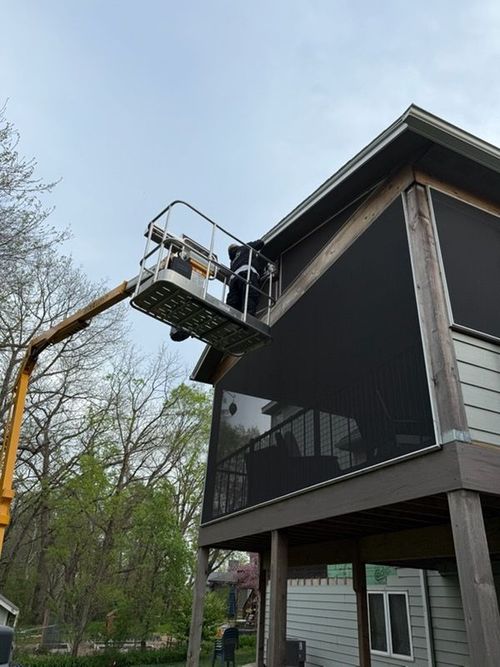 A worker in a bucket lift repairs the roofline of a second-story screened-in porch on a house surrounded by trees.