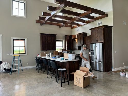 Workers install cabinets in a kitchen with high ceilings, exposed wood beams, and a large island on a polished concrete floor.