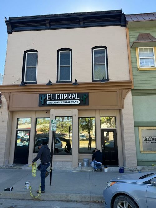 Two people paint the storefront of a tan-colored building labeled