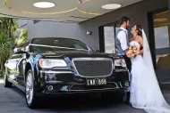 Newlyweds stand beside a black Chrysler limousine. The bride holds a bouquet, and the groom smiles.