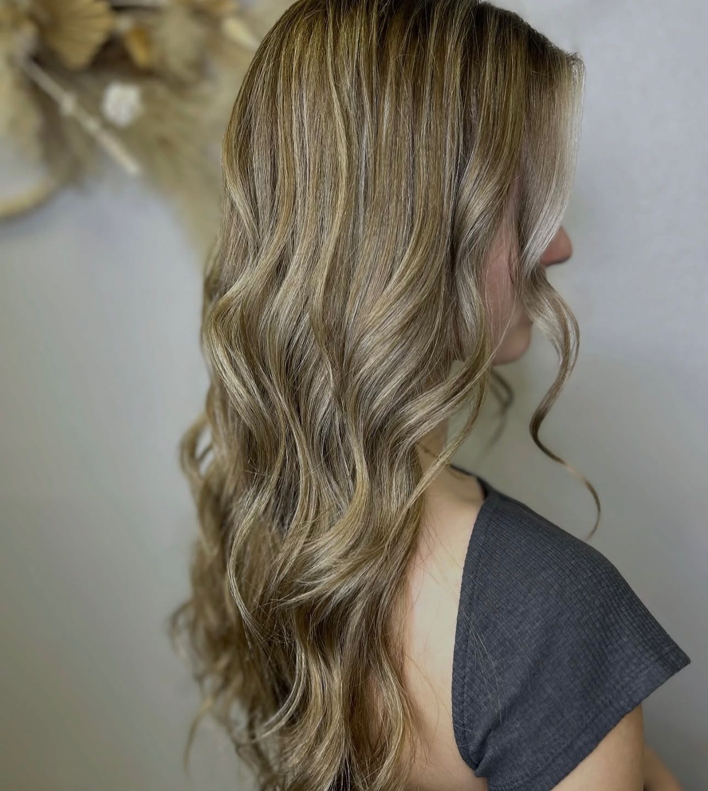 A side view of long, light brown wavy hair with subtle highlights, styled in soft curls against a plain light background.