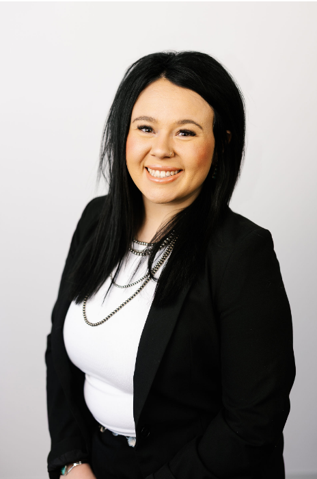 A professional portrait of a person with long dark hair, wearing a white shirt and a black blazer, smiling at the camera.