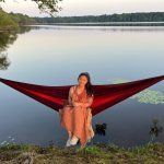 A woman is sitting in a hammock on the shore of a lake.
