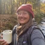 A woman is taking a selfie in the woods while holding a cup of coffee.