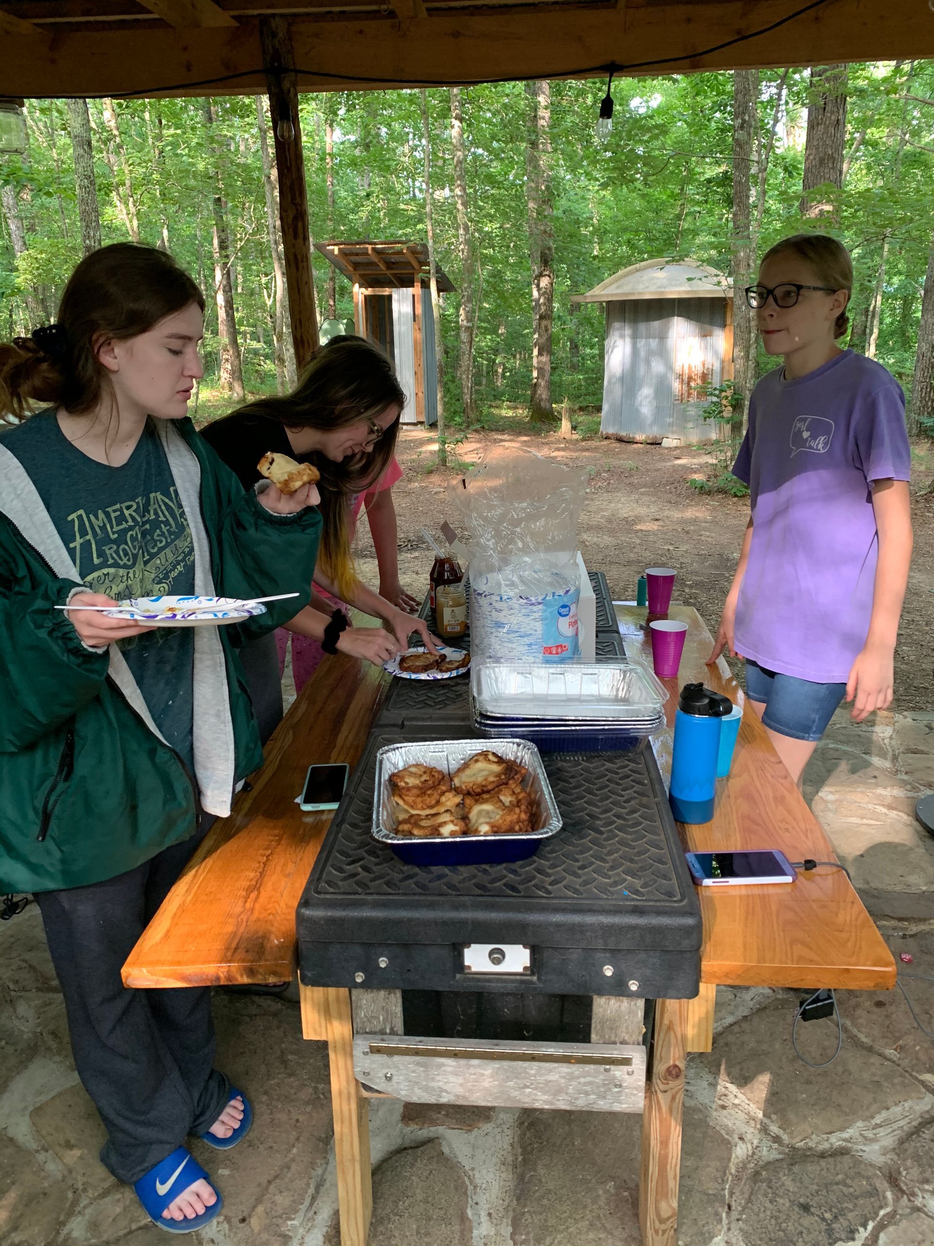 A group of young girls are standing around a picnic table eating food.