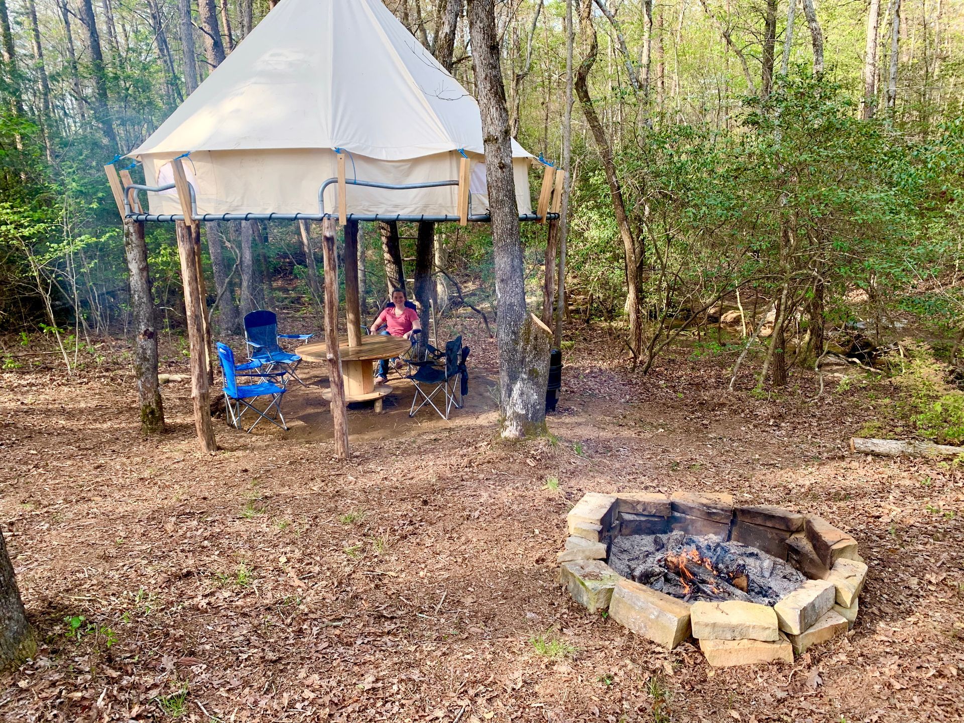 A tent is sitting in the middle of a forest next to a fire pit.