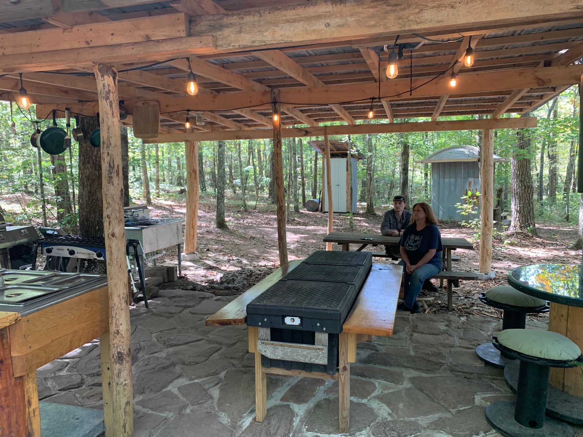 A group of people are sitting at tables under a covered area in the woods.