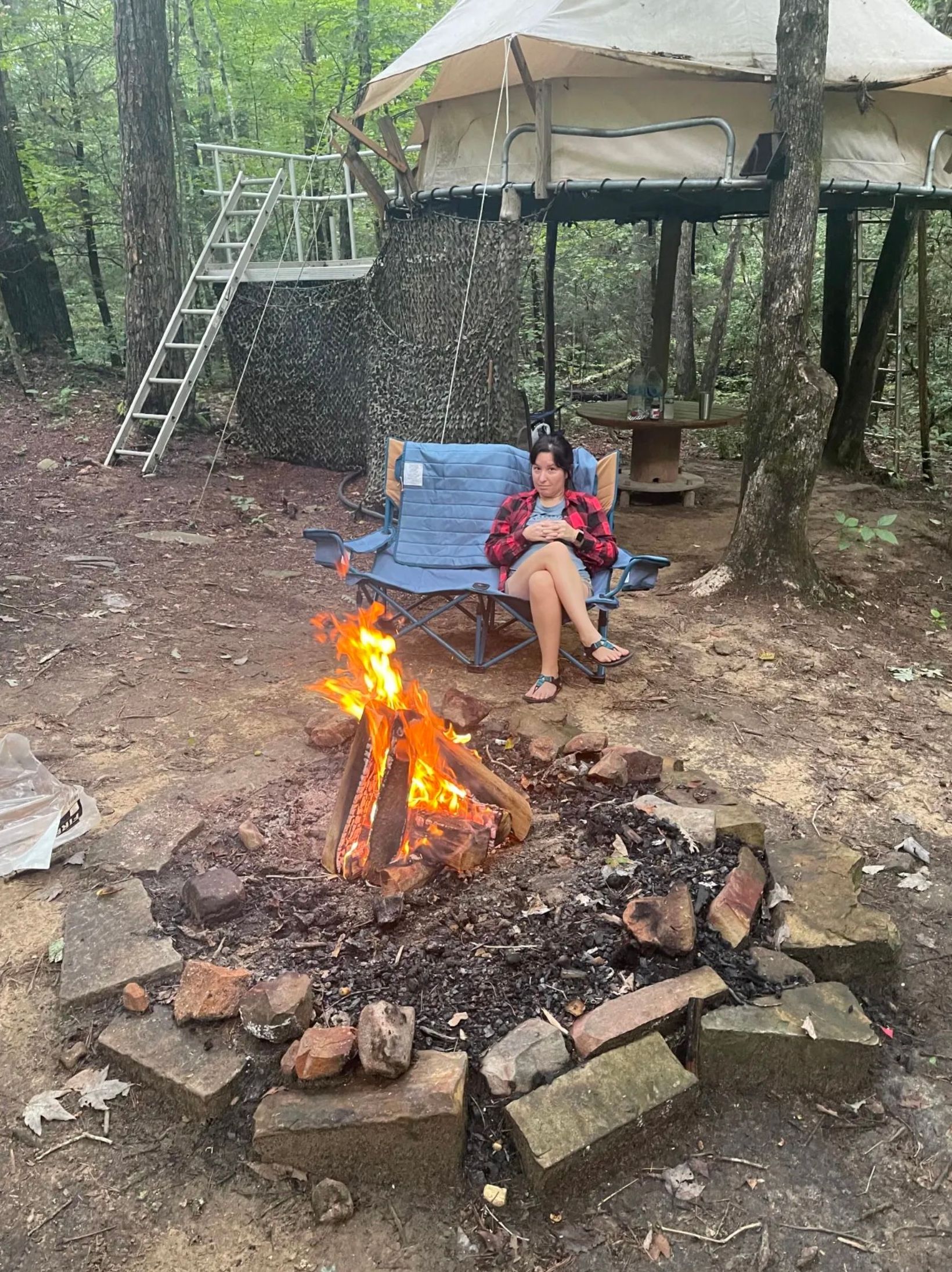 A woman is sitting in a chair next to a campfire in the woods.