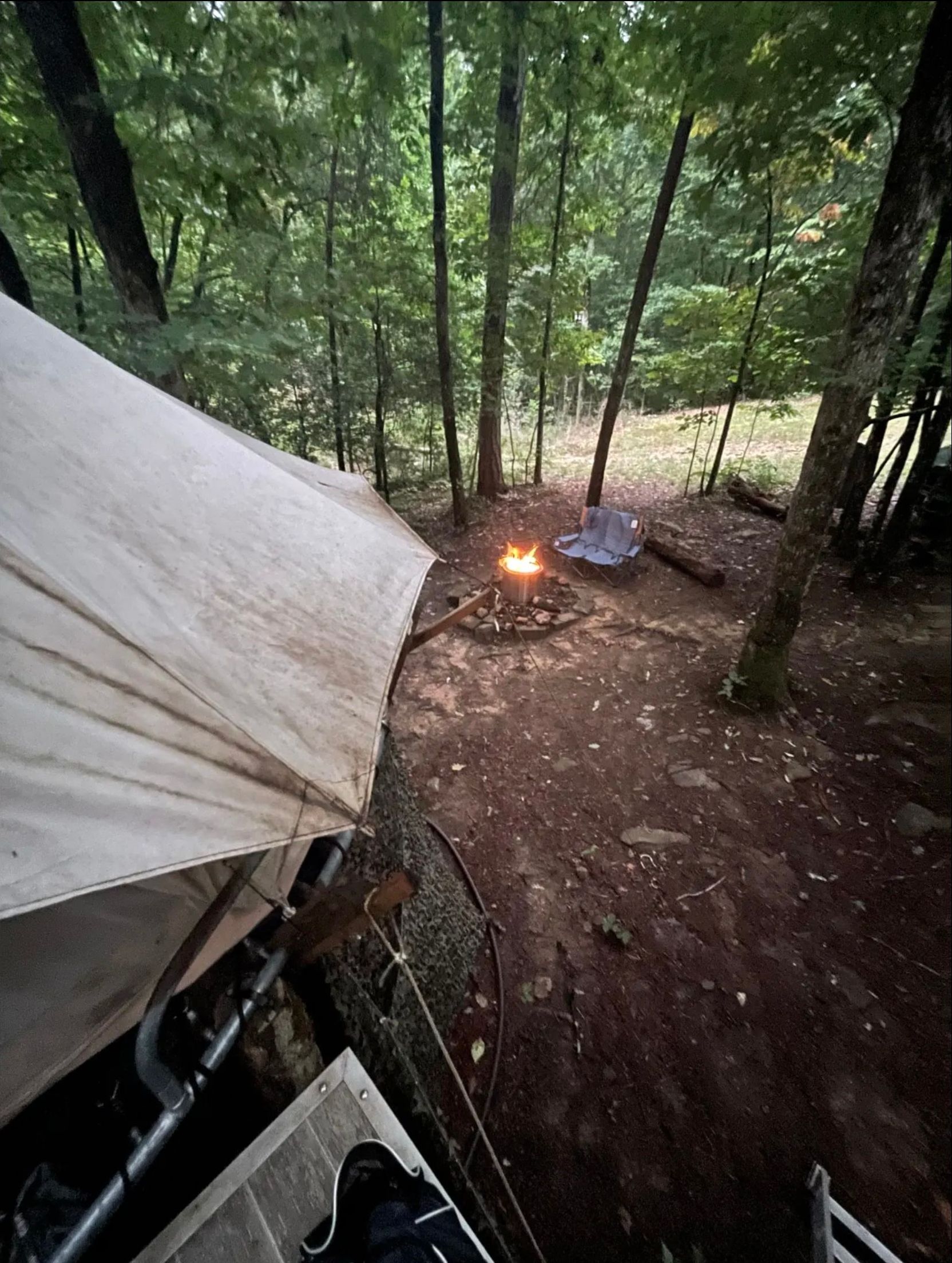 A tent is sitting in the middle of a forest next to a campfire.