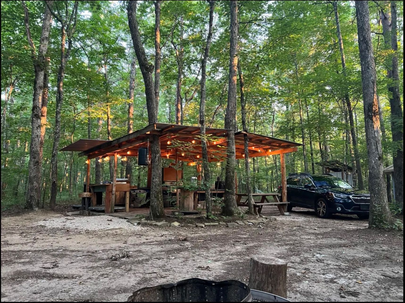 A car is parked in front of a picnic shelter in the woods.