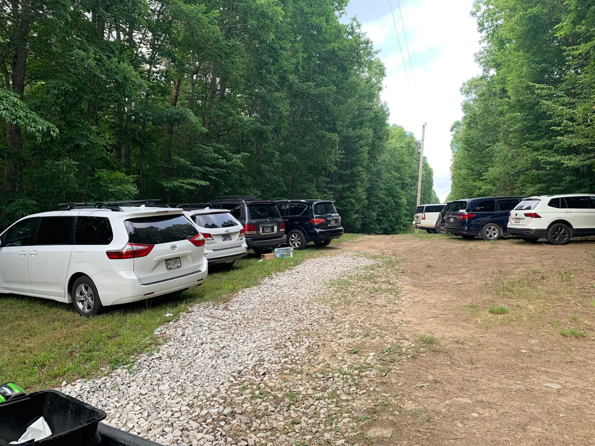 A row of cars are parked in a gravel lot in the woods.