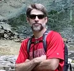 A man wearing sunglasses and a red shirt is standing with his arms crossed.