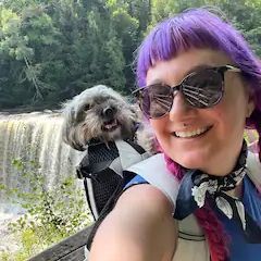 A woman with purple hair is carrying a small dog in a backpack.