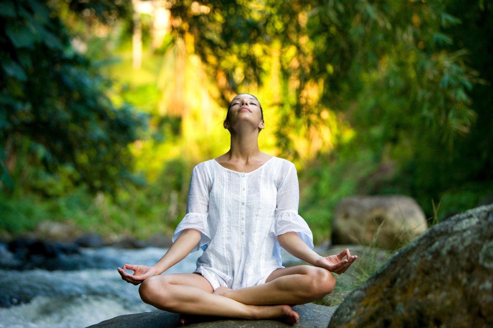 A woman is sitting in a lotus position on a rock by a river.