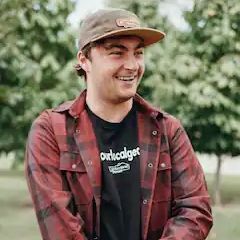 A young man wearing a plaid shirt and a hat is smiling.