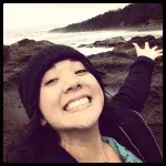 A woman wearing a black hat is smiling and standing on a rocky beach.