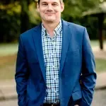 A man in a blue suit and plaid shirt is standing with his hands in his pockets.